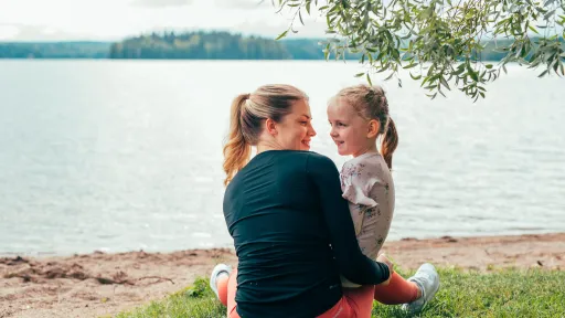 Äiti ja lapsi istuvat nurmikolla järven rannalla kesäisenä päivänä.
