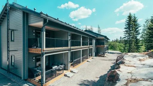 Kaksikerroksinen harmaa puutalo metsämaisemassa, aurinkoinen sää.