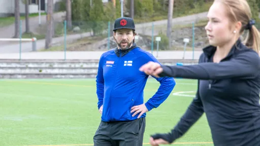 Coach in a blue jacket observing a woman in black running on a field.