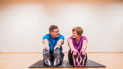 Kaksi henkilöä venyttelee jumppamatolla sisätiloissa, hymyilevät toisilleen.