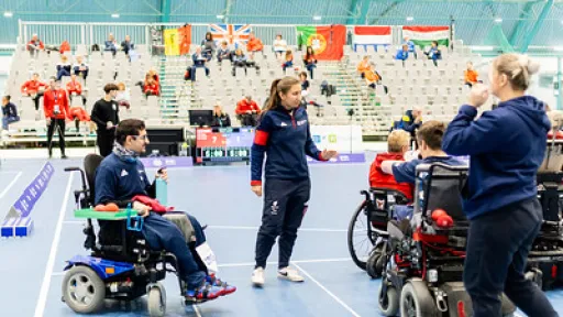 Hallissa peli pyörätuolissa, pelaajat ja valmentaja keskustelevat. Taustalla katsojia.