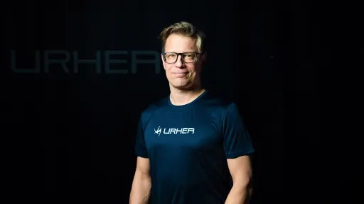 Henkilö seisoo tummassa ympäristössä, päällään sininen paita ja silmälasit.