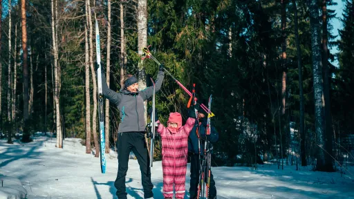Kolme henkilöä hiihtää lumisessa metsässä, iloisia ja nostavat sauvat ylös.