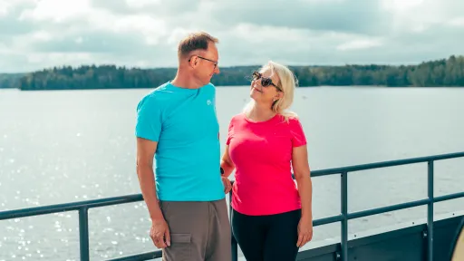 Man and woman smiling at each other on a boat, lake and trees in background.