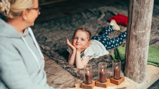 Hymyilevä tyttö makaa laavulla taljan päällä mummon katsoessa vieressä.