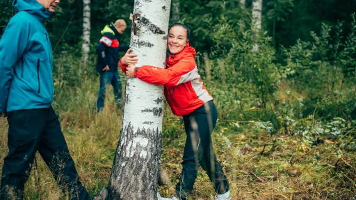 Nainen venyttelee jalkaa puuta vasten, hymyilee ja halaa puun runkoa