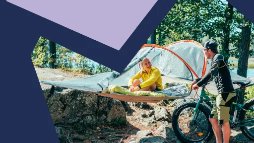 Järven rannalla puihin on kiinnitetty tentsile, jossa nainen istuu ja hymyilee fatbikea pitelevälle miehelle.