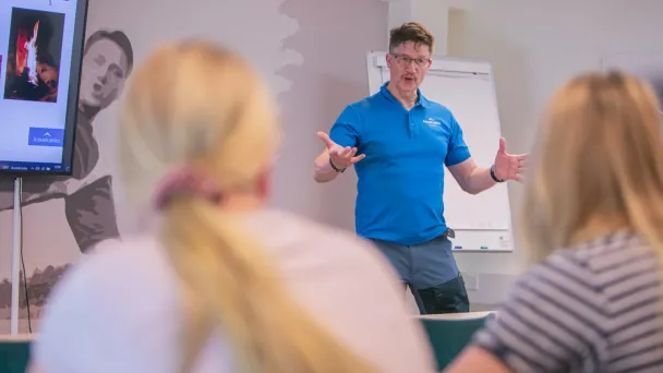 Mies sinisessä paidassa esittelee kahdelle kuuntelijalle luokkahuoneessa.
