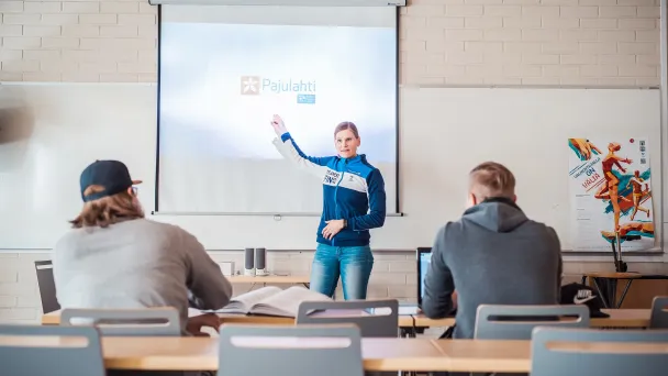 Luokkahuone, opettaja näyttää diaa, opiskelijat istuvat pöydissä.