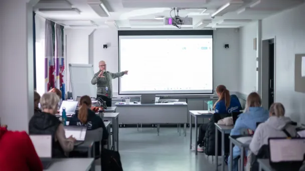 Opettaja luennoi luokassa oppilaille, taustalla valkokangas.