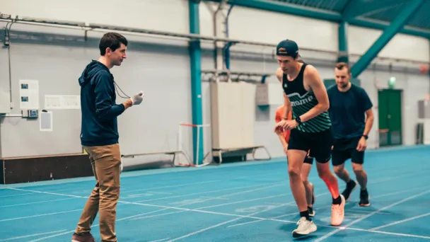 Athletes running on an indoor track while a coach times them.
