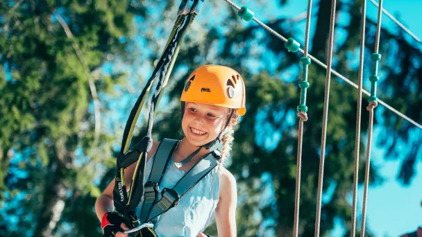 Nuori tyttö kiipeilyvarusteissa hymyilee köysiradalla, aurinkoinen sää ja metsätausta.
