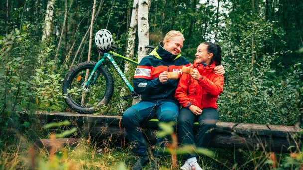 Pari istuu metsässä pyörän kanssa, hymyilee toisilleen.