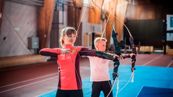 Child and teen practicing archery indoors on a blue mat.