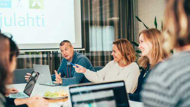 Iloinen ryhmä kokouksessa neuvotteluhuoneessa, kannettavat tietokoneet pöydällä.