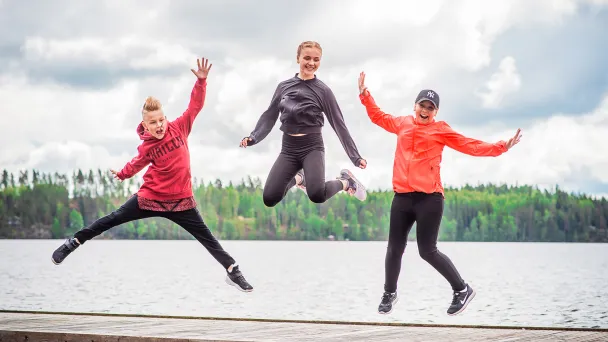 Kolme ihmistä hyppää laiturilla järven edessä, taustalla metsää.