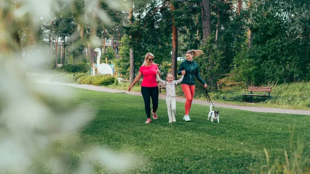 Pajulahden rantanurmella iloinen lapsi pitää kädestä mummoa ja äitiä koiran kävellessä vierellä.