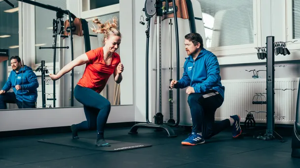 Woman exercising indoors while trainer observes.