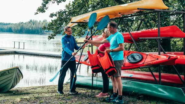 Three people by kayaks at a lakeside, holding paddles and life jackets.