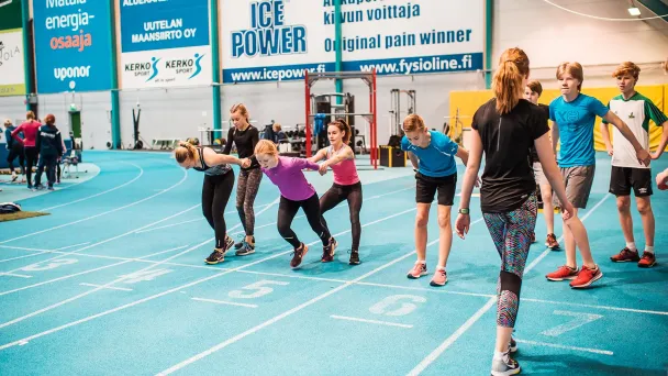 Indoor track practice with young athletes lined up and a coach giving instructions.