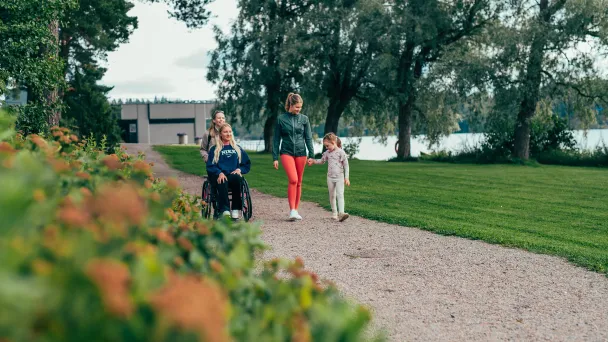Kolme ihmistä kävelyllä puistossa järven rannalla, yksi henkilö pyörätuolissa.