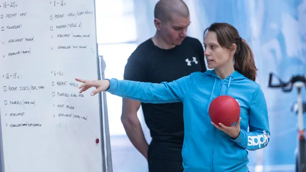 Naishenkilö sinisessä hupparissa osoittaa taululle, pallo kädessä.