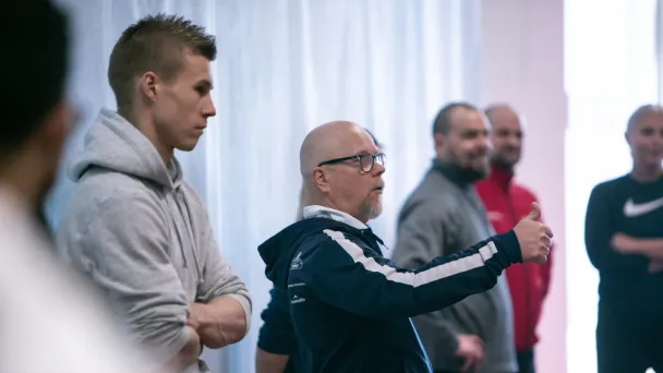 Useat miehet seisovat keskustellen sisätiloissa, yksi elehtii kädellään.