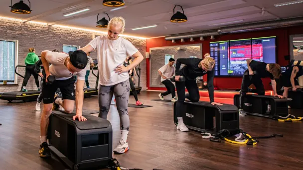 Kuntosalilla ryhmä ihmisiä treenaamassa, taustalla suuret näytöt.