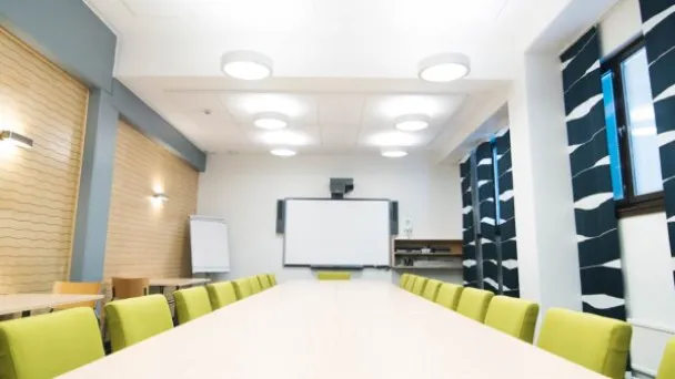 Kokoushuone, pitkät pöydät ja keltaiset tuolit, valkoinen taulu edessä.