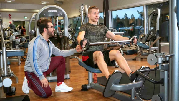 Kuntosalilla henkilö käyttää soutulaitetta ohjaajan avustamana.