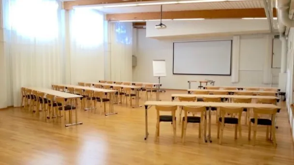Tyhjä luokkahuone puulattialla, pöydät ja tuolit siistissä järjestyksessä.