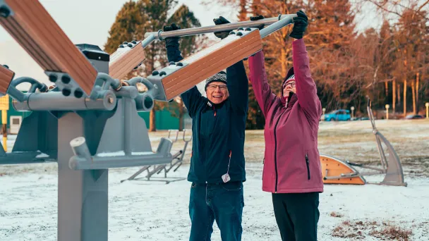 Senioripariskunta kuntoilee Pajulahden rannassa ulkokuntoilulaitteilla hymyillen.