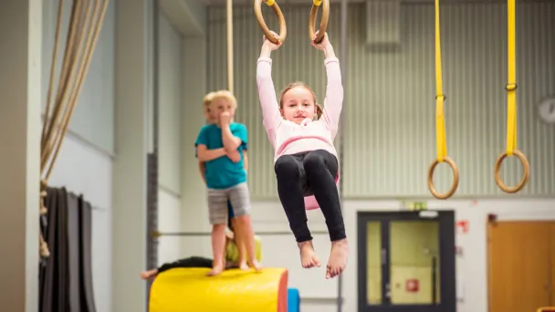 Lapsi roikkuu voimistelurenkaissa, toinen seisoo keltaisella matolla.