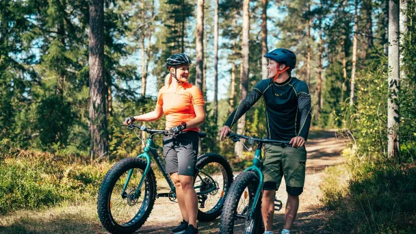 Kaksi henkilöä pyöräilemässä metsäpolulla, aurinkoinen päivä.
