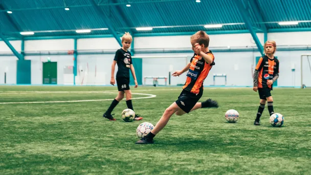 Kolme lasta pelaa jalkapalloa sisätiloissa sinisellä kentällä.