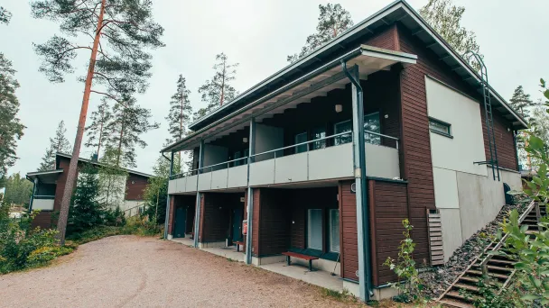 Kaksikerroksinen puutalo metsän keskellä, ympärillä hiekkaa ja puita.