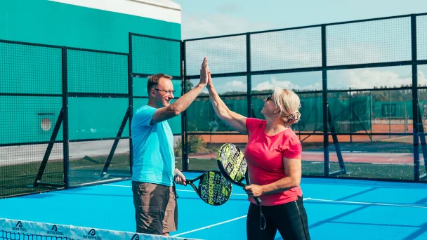 Kaksi henkilöä pelaa padelia ja antaa ylävitosta kentällä.