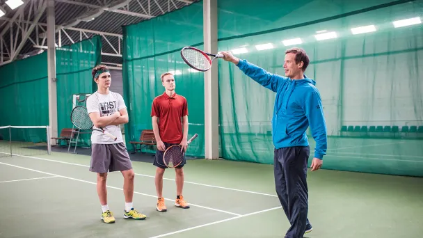 Tennisharjoitus hallissa, valmentaja ohjaa kahta nuorta pelaajaa.