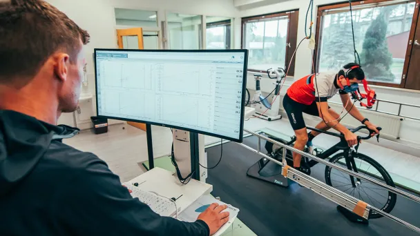 Sisätiloissa pyöräilevä henkilö testissä, taustalla näyttö ja analysoiva tutkija.