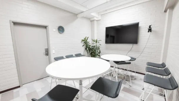 Tyylikäs kokoushuone, kaksi pyöreää pöytää, tuoleja ja seinällä televisio.