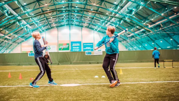 Kaksi henkilöä sisäjalkapallohallissa, harjoittelevat pesäpallon lyömistä.