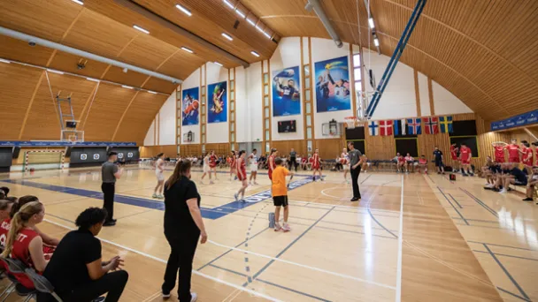 Koripallo-ottelu liikuntasalissa, pelaajat ja tuomarit toiminnassa.