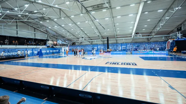 Koripallokenttä sisähallissa, katsojia taustalla.