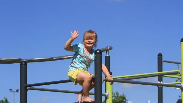 Child smiling and waving on playground bars under a clear blue sky.