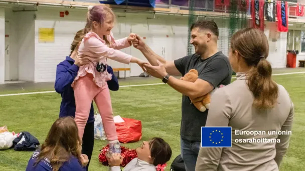 Aikuiset auttavat hymyilevää lasta seisomaan ulkonaurheilutilassa.