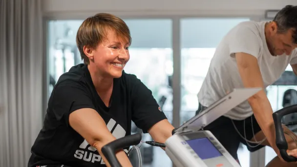 Hymyilevä nainen kuntopyörällä valoisassa huoneessa, vierellään toinen pyöräilijä.