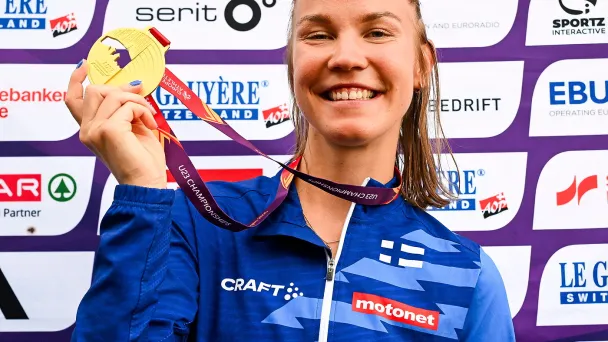 Tähtiurheilija sinisessä asussa, hymyilee ja pitää kultaista mitalia taustallaan sponsoreiden logoja.