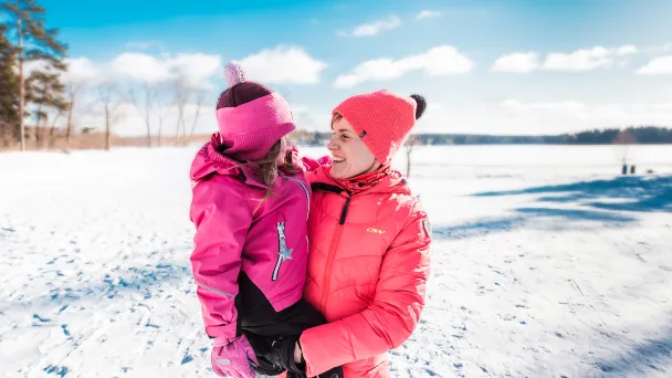 Hymyilevä nainen pitelee sylissään lasta, taustalla kaunis, talvinen järvimaisema.