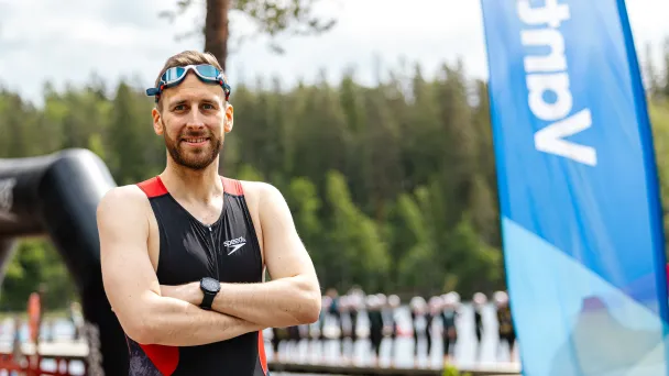 Mies triathlon-puvussa seisoo kädet ristissä, taustalla järvimaisema ja banneri.