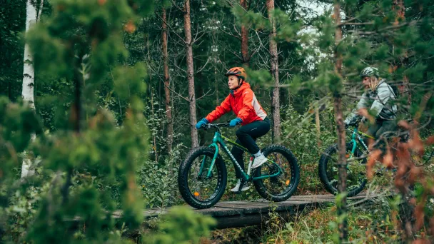 Kaksi ihmistä pyöräilee fatbikeilla metsässä ojan yli menevän sillan päällä.